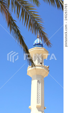 Spire Rises Amidst Leafy Fronds. Tall Spire Peers Beyond Swaying Palm Leaves Beneath Blue Sky. Majestic Minaret Ascends High Above Vibrant And Gentle Tropical Landscape 140263765