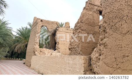 Weathered Adobe Meets Lively Foliage. Fragile Wall Near Lush Palm Grove Reveals History. Desert Border Showcases Historic Walls Against Thriving Green Surroundings Weathered Adobe Meets Lively Foliage. Fragile Wall Near Lush Palm Grove Reveals History. Desert Border Showcases Historic Walls Against Thriving Green Surroundings 140263782