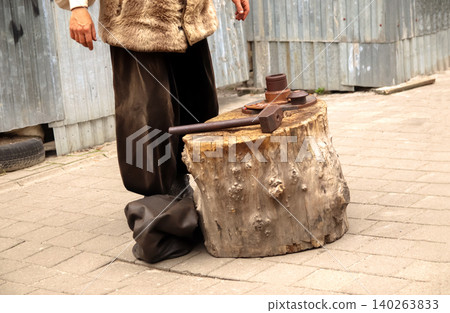 Cropped shot of a man in traditional Ukrainian Cossack clothes standing near a wooden anvil stump with a hammer. Historical reenactment of old metal blacksmith or coin minting craft Cropped shot of a man in traditional Ukrainian Cossack clothes standing near a wooden anvil stump with a hammer. Historical reenactment of old metal blacksmith or coin minting craft 140263833