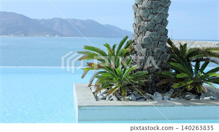 Calm Coastal Retreat. Tranquil Infinity Edge Pool Overlooking Calm Azure Ocean And Mountains. Serene Sunlit Terrace With Lush Palms And Smooth Tiles Over Peaceful Sea Scene 140263835
