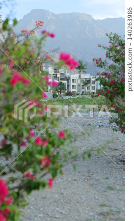 Calm Pathway Surrounded By Lush Greenery Leading To Seaside Villas. Idyllic Gravel Route With Blooming Flora Guiding Visitors To Coastal Retreats Under Bright Sunny Sky Calm Pathway Surrounded By Lush Greenery Leading To Seaside Villas. Idyllic Gravel Route With Blooming Flora Guiding Visitors To Coastal Retreats Under Bright Sunny Sky 140263986