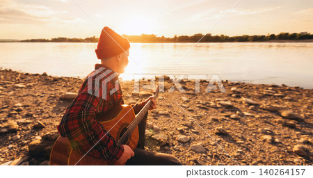 Musician Playing Acoustic Guitar by River at Sunset 140264157
