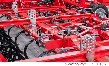 Red metal farm implement with multiple wheels, brackets, and curved tines arranged in rows, shown indoors with a clean industrial background and bright reflective surfaces Red metal farm implement with multiple wheels, brackets, and curved tines arranged in rows, shown indoors with a clean industrial background and bright reflective surfaces 140264307