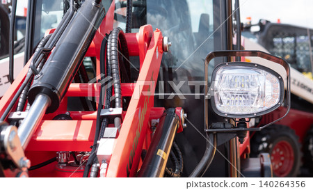 Detailed close-up of red farm machinery showing hydraulic cylinders, metal arms, hoses, and a front work light beside the cab, with industrial equipment visible in the background Detailed close-up of red farm machinery showing hydraulic cylinders, metal arms, hoses, and a front work light beside the cab, with industrial equipment visible in the background 140264356