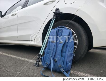 Blue backpack and trekking poles leaning against a white compact car parked on asphalt. High quality photo 140264457