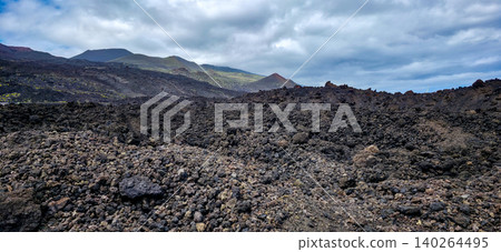 Volcanic landscape featuring rugged black lava rock formations with distant green hills and cloudy sky in the background, showcasing the aftermath of volcanic activity. High quality photo Volcanic landscape featuring rugged black lava rock formations with distant green hills and cloudy sky in the background, showcasing the aftermath of volcanic activity. High quality photo 140264495