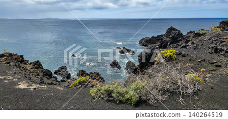 Rocky coastal landscape with black volcanic rocks, green vegetation, and calm ocean waters under a cloudy sky in a scenic natural environment. High quality photo Rocky coastal landscape with black volcanic rocks, green vegetation, and calm ocean waters under a cloudy sky in a scenic natural environment. High quality photo 140264519