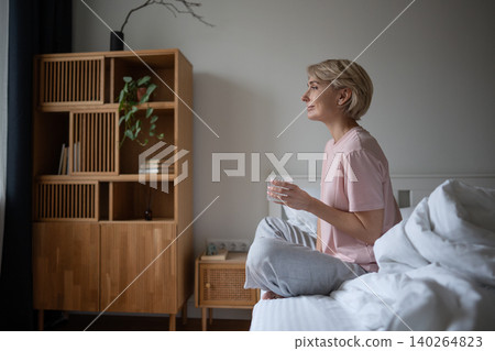 Thoughtful positive woman sitting in bed in bedroom with glass of water peacefully looking at window 140264823