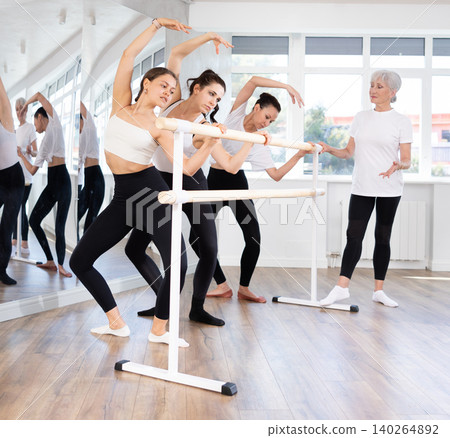 Female ballet trainer teaching group of dancers near ballet barre in dancing studio Female ballet trainer teaching group of dancers near ballet barre in dancing studio 140264892