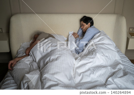 Sleepless elderly female lying in bed while husband enjoying night sleep and snoring loudly Sleepless elderly female lying in bed while husband enjoying night sleep and snoring loudly 140264894