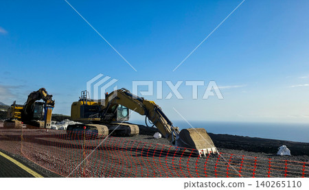 Specialized equipment machine with hydraulic arm positioned on volcanic rock and solidified lava overlooking the ocean under a clear blue sky with distant mountains visible. High quality photo Specialized equipment machine with hydraulic arm positioned on volcanic rock and solidified lava overlooking the ocean under a clear blue sky with distant mountains visible. High quality photo 140265110