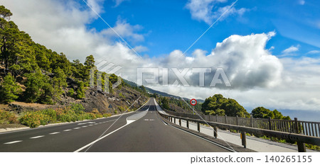Scenic winding road through lush green hills under a bright blue sky with fluffy white clouds, showcasing a tranquil landscape in a mountainous volcanic region. High quality photo Scenic winding road through lush green hills under a bright blue sky with fluffy white clouds, showcasing a tranquil landscape in a mountainous volcanic region. High quality photo 140265155