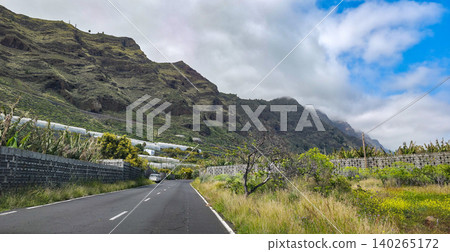 Scenic road winding through lush banana plantations with steep mountainous terrain under a partly cloudy sky in a tropical landscape. High quality photo 140265172