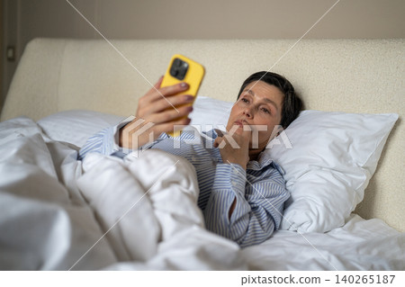 Thoughtful elderly woman in bed procrastinating watching news online on smartphone after waking up. Thoughtful elderly woman in bed procrastinating watching news online on smartphone after waking up. 140265187
