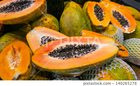 Freshly cut papaya fruit displayed among whole papayas in a grocery store, showcasing vibrant orange flesh and black seeds, with protective netting visible. High quality photo 140265278