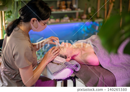 Relaxed woman receiving traditional exotic Gua Sha facial massage therapy with stone scrubber. Relaxed woman receiving traditional exotic Gua Sha facial massage therapy with stone scrubber. 140265380
