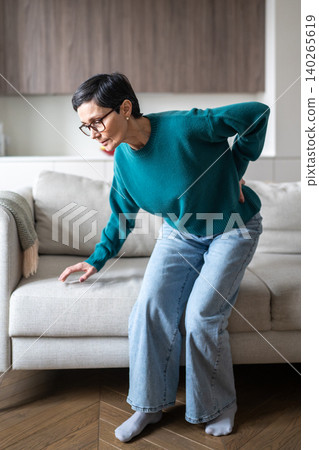 Senior woman with spinal disorder suffering from sudden back pain after rising up from sofa. Senior woman with spinal disorder suffering from sudden back pain after rising up from sofa. 140265619