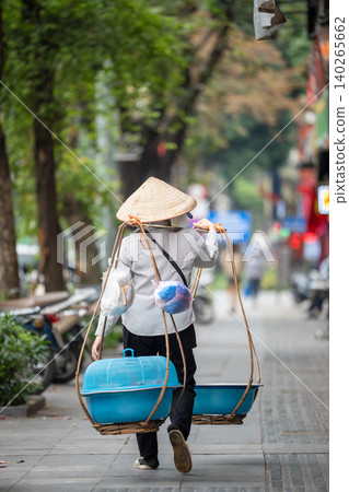 Unrecognizable street vendor person in conical hat walking down street in Hanoi carrying stuff on shoulder pole. Unrecognizable street vendor person in conical hat walking down street in Hanoi carrying stuff on shoulder pole. 140265662