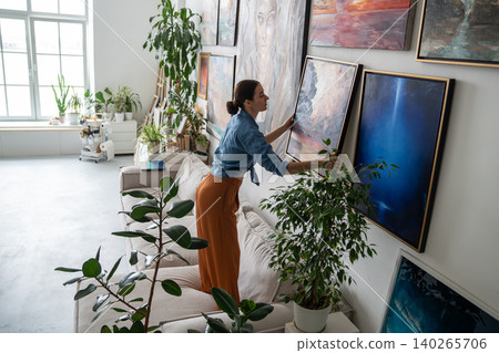 Talented woman painter hanging new finished artwork canvas on wall at home loft gallery studio. Talented woman painter hanging new finished artwork canvas on wall at home loft gallery studio. 140265706