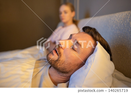Man sleeping in bed and snoring loud while frustrated wife sitting beside with exhausted expression. Man sleeping in bed and snoring loud while frustrated wife sitting beside with exhausted expression. 140265719