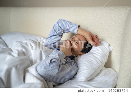 Dreamy senior woman comfortably lying in bed covered in cozy blanket and yawning after waking up. Dreamy senior woman comfortably lying in bed covered in cozy blanket and yawning after waking up. 140265734