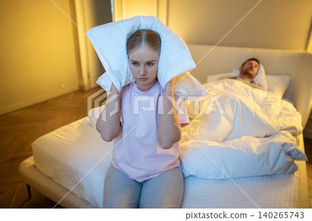 Irritated wife sitting on bed covering ears with pillow to block snore sound while man sleeping. Irritated wife sitting on bed covering ears with pillow to block snore sound while man sleeping. 140265743