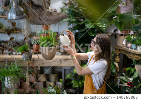 Woman gardener in overalls watering potted houseplant in greenhouse surrounded by plants and pots Woman gardener in overalls watering potted houseplant in greenhouse surrounded by plants and pots 140265868