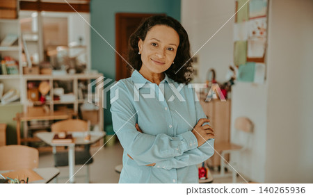 Warm mature Hispanic teacher smiling with arms crossed in a bright elementary classroom Warm mature Hispanic teacher smiling with arms crossed in a bright elementary classroom 140265936