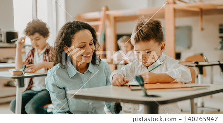 Hispanic female teacher helping young Caucasian schoolboy writing at desk in classroom Hispanic female teacher helping young Caucasian schoolboy writing at desk in classroom 140265947
