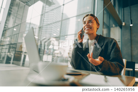 Happy young adult Caucasian woman talking on smartphone in a modern city cafe near laptop Happy young adult Caucasian woman talking on smartphone in a modern city cafe near laptop 140265956