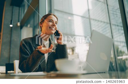 Happy young Caucasian woman talking on phone and working with laptop inside modern cafe Happy young Caucasian woman talking on phone and working with laptop inside modern cafe 140265963