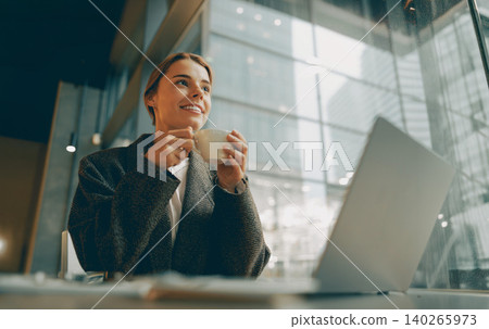 Smiling young adult Caucasian woman drinking coffee near laptop in a modern city cafe Smiling young adult Caucasian woman drinking coffee near laptop in a modern city cafe 140265973