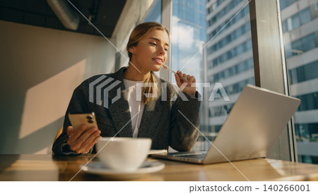 Thoughtful young Caucasian woman working on laptop and holding phone in modern city cafe Thoughtful young Caucasian woman working on laptop and holding phone in modern city cafe 140266001