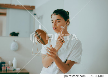 Young Caucasian woman making playful face while using rose quartz roller in bright bathroom Young Caucasian woman making playful face while using rose quartz roller in bright bathroom 140266009