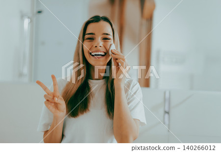 Happy young adult Caucasian woman cleanses face and makes peace sign in bright bathroom Happy young adult Caucasian woman cleanses face and makes peace sign in bright bathroom 140266012