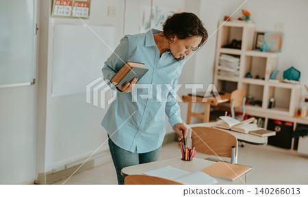 Middle aged Latina teacher holding book and arranging pens on desk in classroom Middle aged Latina teacher holding book and arranging pens on desk in classroom 140266013