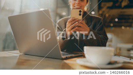 Caucasian young adult woman using her smartphone next to a laptop inside a warm cafe 140266016