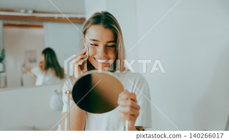 Happy young Caucasian woman cleaning face with cotton pad looking in hand mirror at home Happy young Caucasian woman cleaning face with cotton pad looking in hand mirror at home 140266017