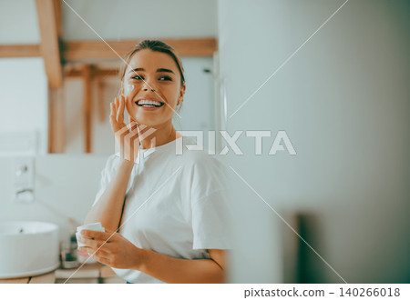 Happy young Caucasian woman in her twenties applying skin cream in a bright bathroom Happy young Caucasian woman in her twenties applying skin cream in a bright bathroom 140266018