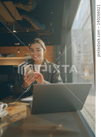 Happy young Caucasian woman using smartphone and working on laptop inside a sunny cafe Happy young Caucasian woman using smartphone and working on laptop inside a sunny cafe 140266021