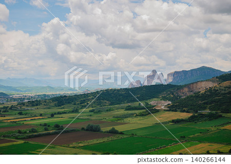 Scenic view of vibrant green agricultural fields and rural meadows in the Thessaly valley with majestic Meteora rocky mountains under a cloudy sky in Greece Scenic view of vibrant green agricultural fields and rural meadows in the Thessaly valley with majestic Meteora rocky mountains under a cloudy sky in Greece 140266144