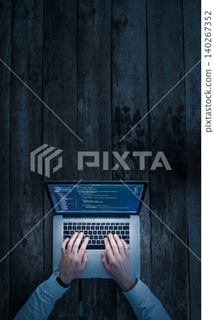 Person works on laptop with code on screen while sitting at wooden table in dim light 140267352