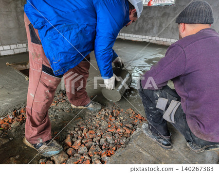 Construction workers pouring fresh wet concrete mix from a bucket onto a floor in an unfinished building, creating a new solid surface during home renovation project. Construction workers pouring fresh wet concrete mix from a bucket onto a floor in an unfinished building, creating a new solid surface during home renovation project. 140267383