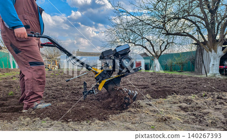 Man using a motor cultivator to till brown garden soil, preparing the ground for planting, symbolizing hard work, spring farming, and sustainable agriculture. 140267393