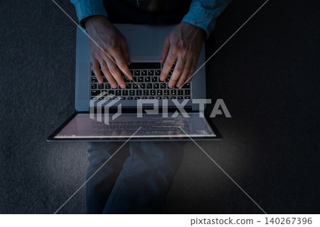 Person types on laptop while seated in a dimly lit room during evening hours 140267396