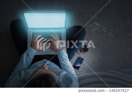 Person works on laptop while sitting on floor in evening with drink beside phone Person works on laptop while sitting on floor in evening with drink beside phone 140267427