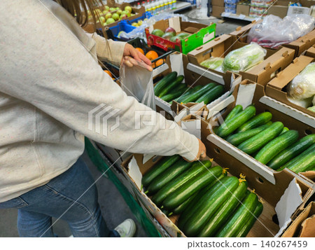 Woman's hand reaching into a cardboard crate of fresh green cucumbers at a supermarket produce aisle, selecting healthy ingredients for cooking and a plant-based diet. 140267459