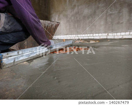 Construction worker in protective gloves leveling freshly poured concrete screed on a building site, ensuring a smooth and even surface for the new floor installation. 140267479