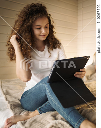 Young woman engaged with tablet while sitting on bed in cozy room during daytime 140267591