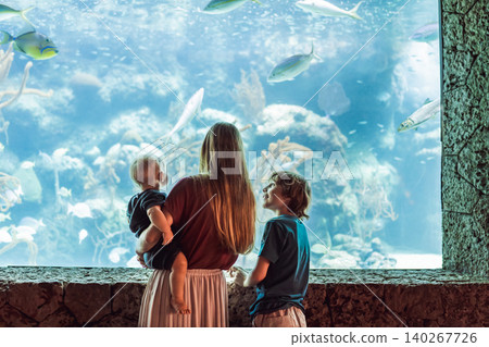 Family sits together by a bright marine tank, showing ocean education, travel with kids, sibling curiosity and underwater wildlife concept 140267726